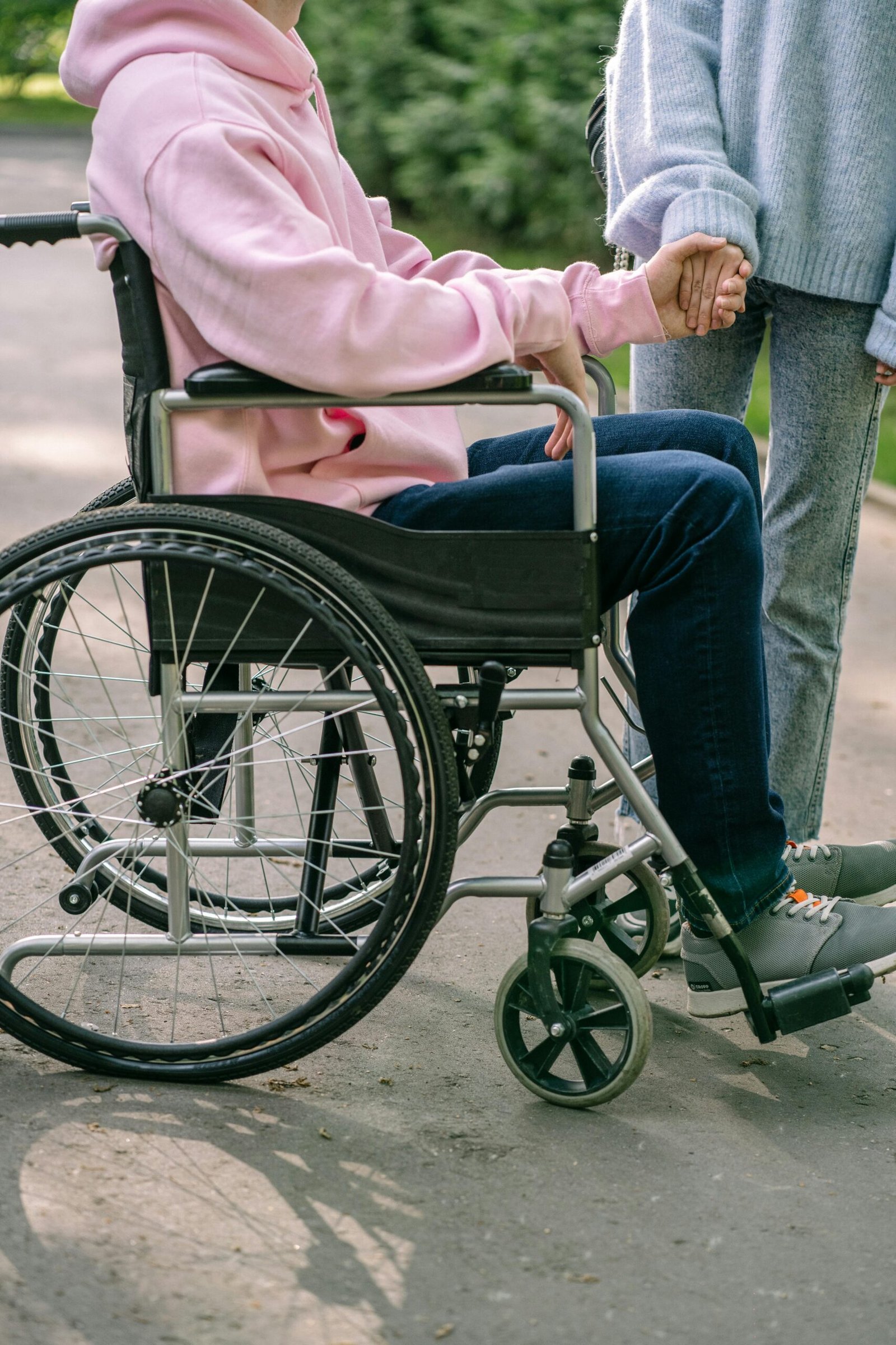 Person in wheelchair holding hands with another individual, showing support and compassion outdoors.
